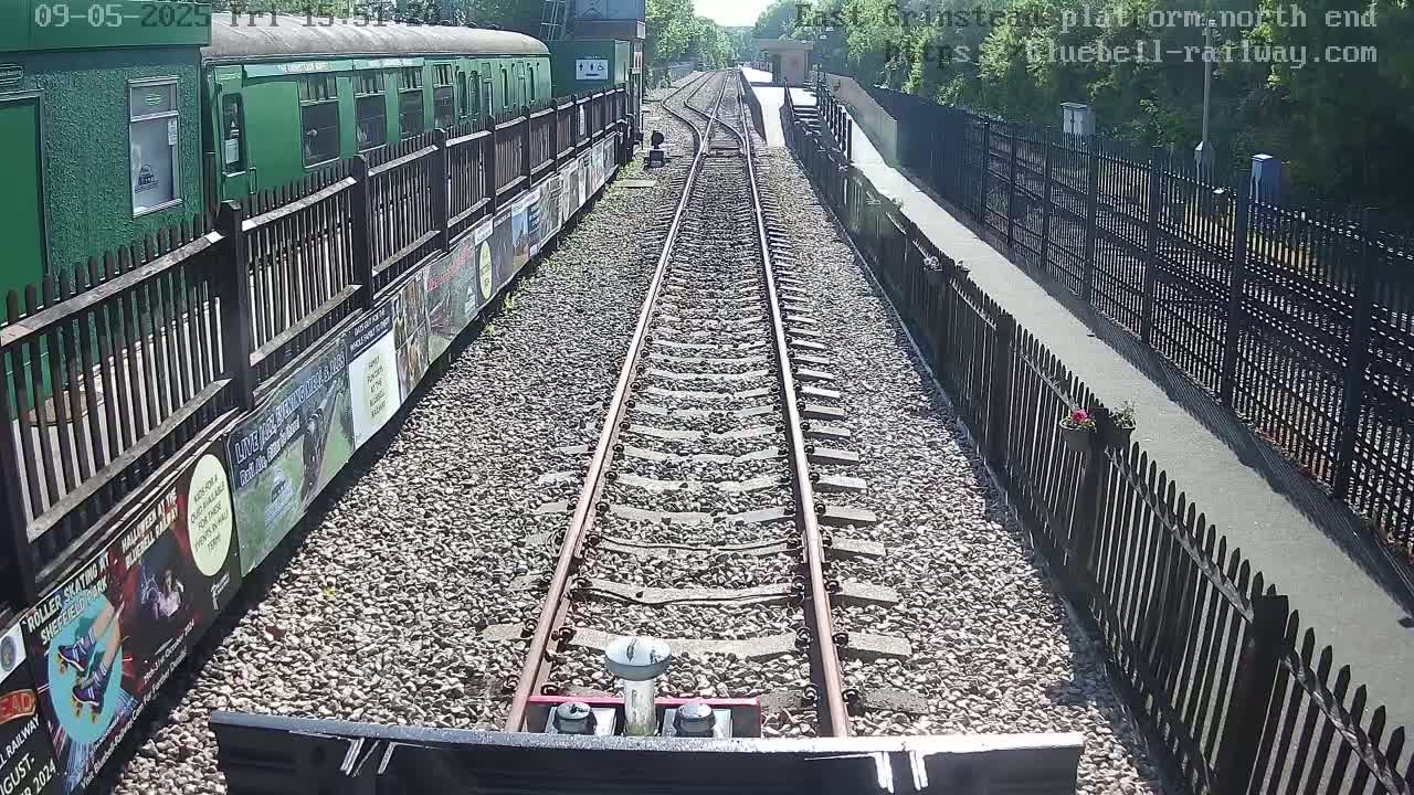 A train travels down a sunny railway track, past a green train car and a fence bordering a paved walkway.
