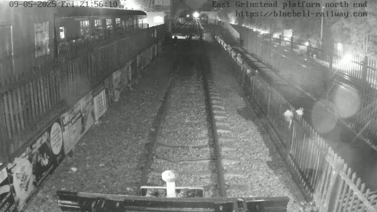 A nighttime view shows a train on a track between fences, with posters on the side of the platform.