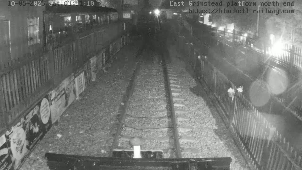 A nighttime, black-and-white view of train tracks extending into a dark tunnel, flanked by fences and posters.