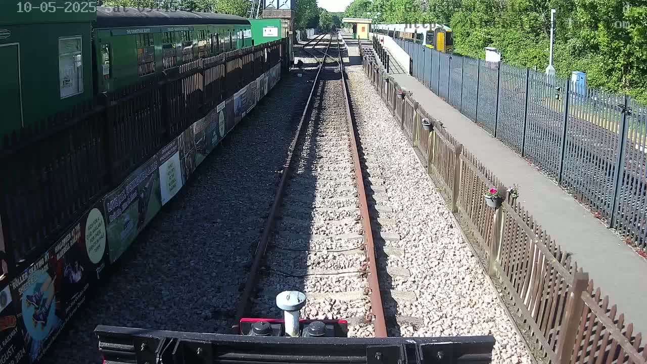 A train is viewed from the front of another train on a sunny day, with railway tracks extending into the distance between fences and train cars.