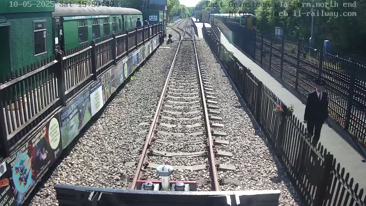 A view from a train shows a railway track extending into the distance on a sunny day, with a green train to the left and a man in a suit walking along a paved path beside a fence to the right.