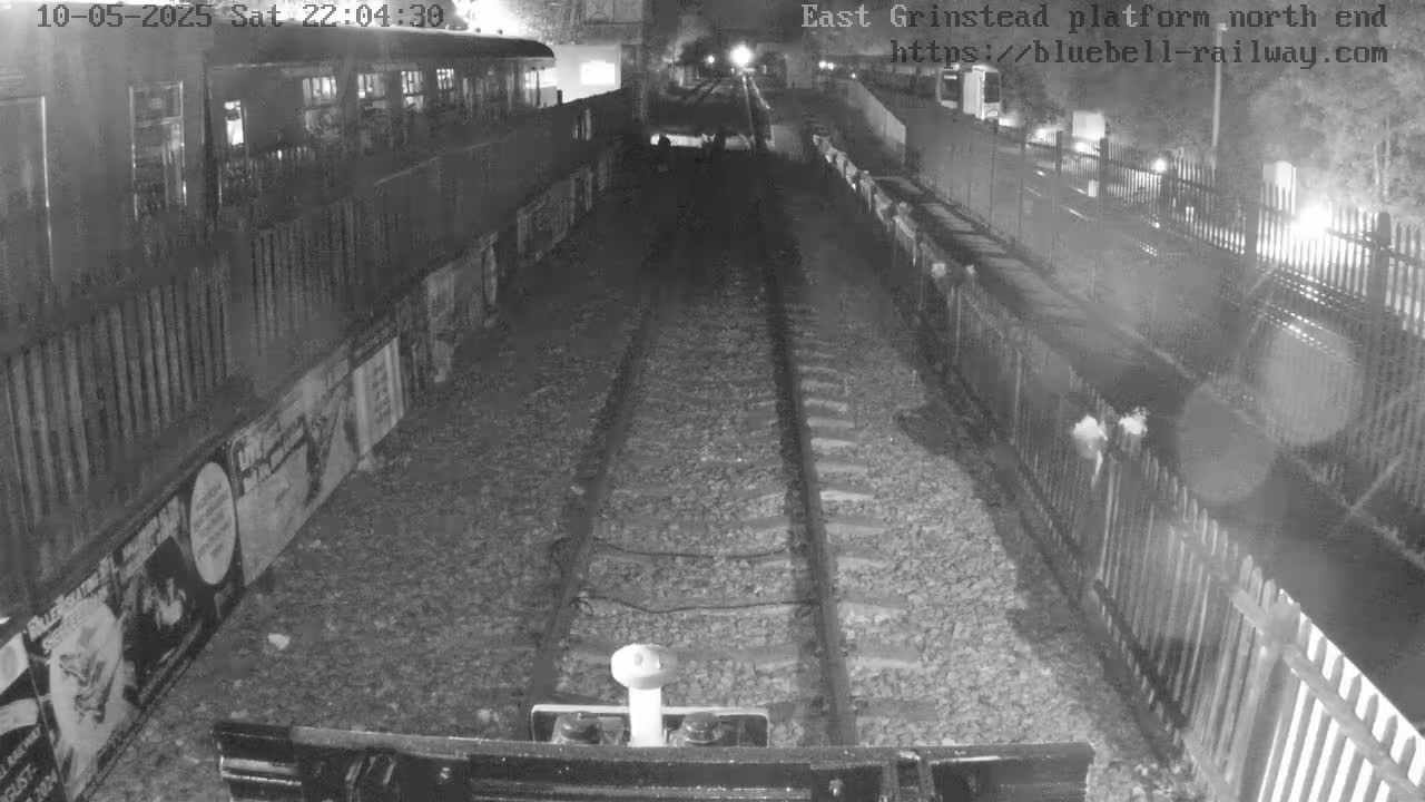 A nighttime view down a train track, bordered by fences and posters, showing a train car to the left and the track extending to a distant light source.