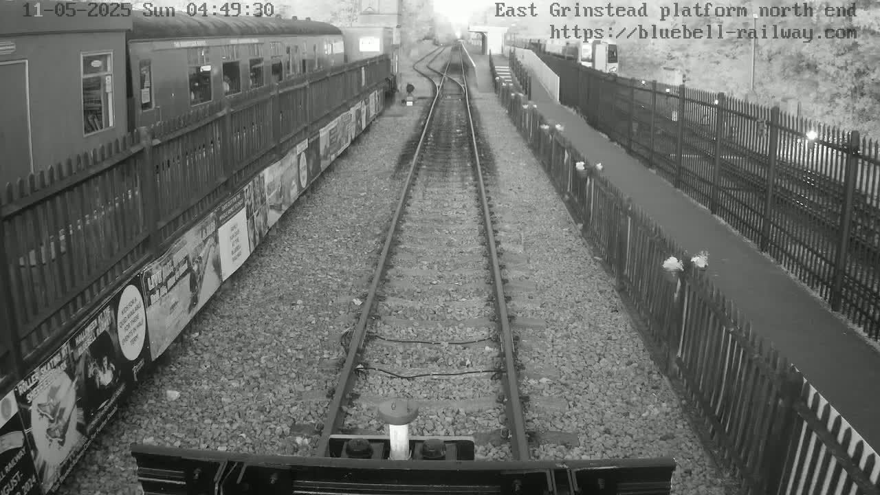A black and white view from a train shows railway tracks extending into the distance between fences, alongside a train car with posters on its side.