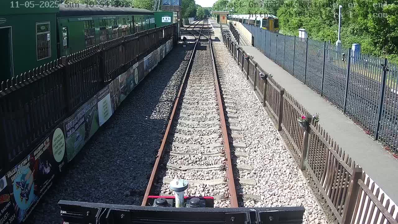 A train track viewed from a low vantage point on a sunny day, bordered by fences and lined with parked trains.