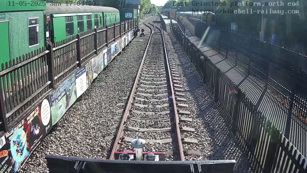 A view down a railway track on a sunny day, with a green train and a fence lined with posters visible on either side.