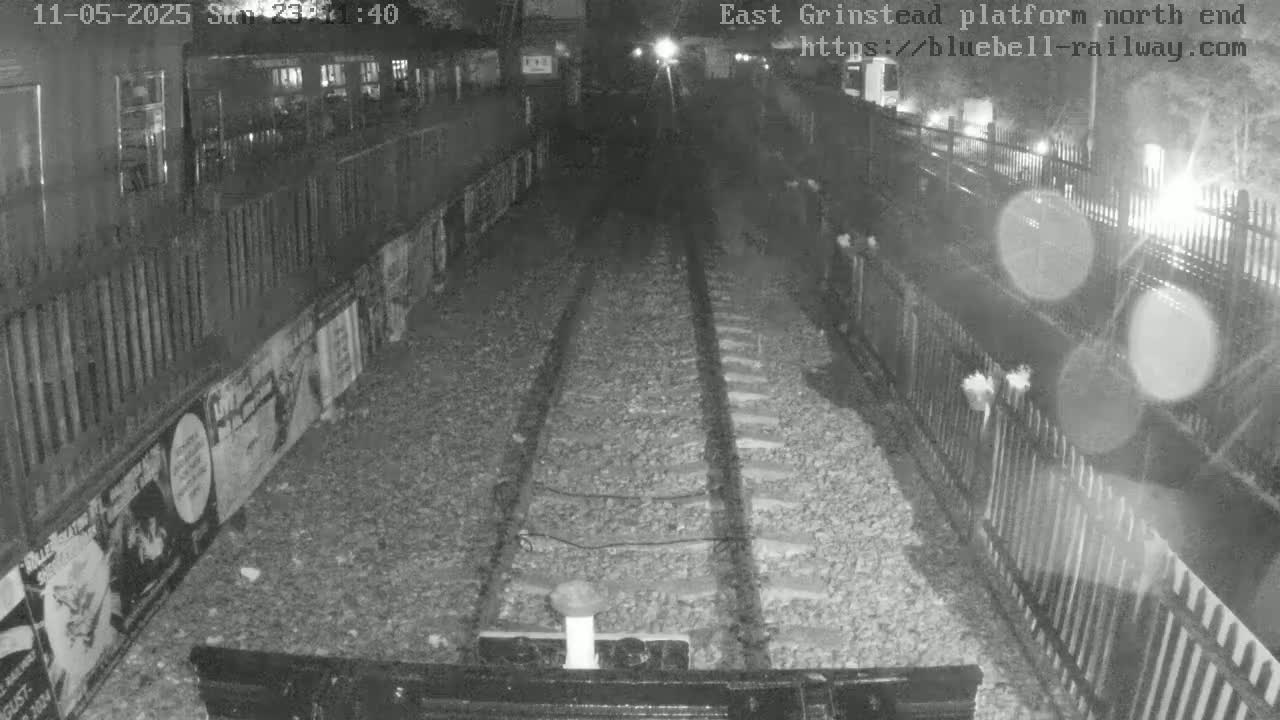 A nighttime view from a train shows railway tracks extending into a dimly lit tunnel, flanked by fences and posters.