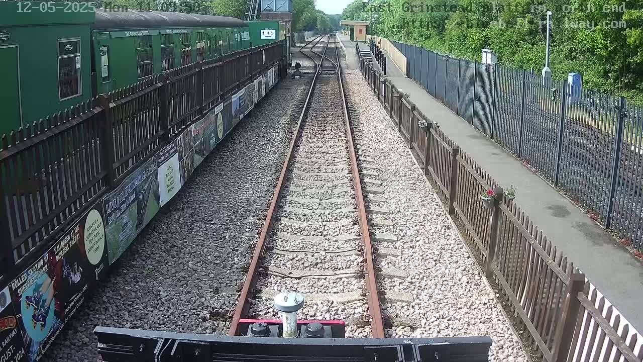 A train's front view shows railway tracks extending toward a green train car at a station on a sunny day.