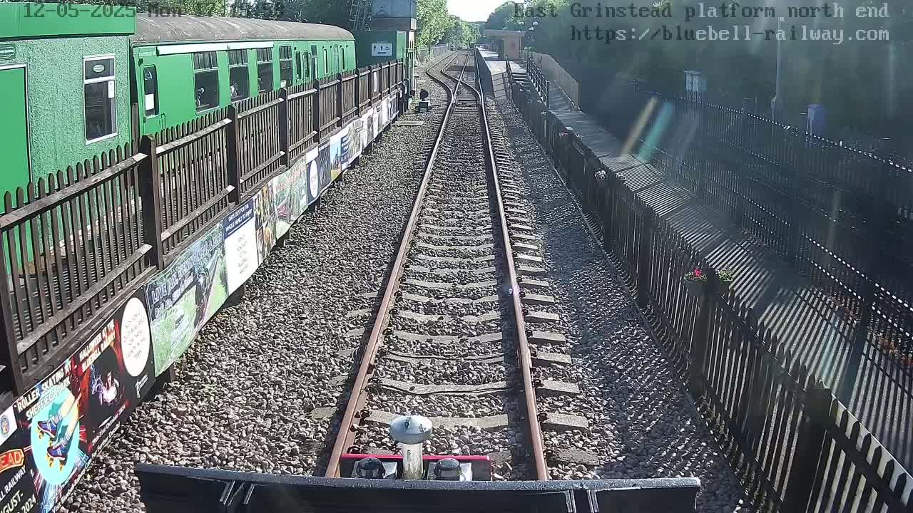 A view from a train shows a sunny day at a train station with a green train alongside a gravel path bordered by fences, and railway tracks extending into the distance.