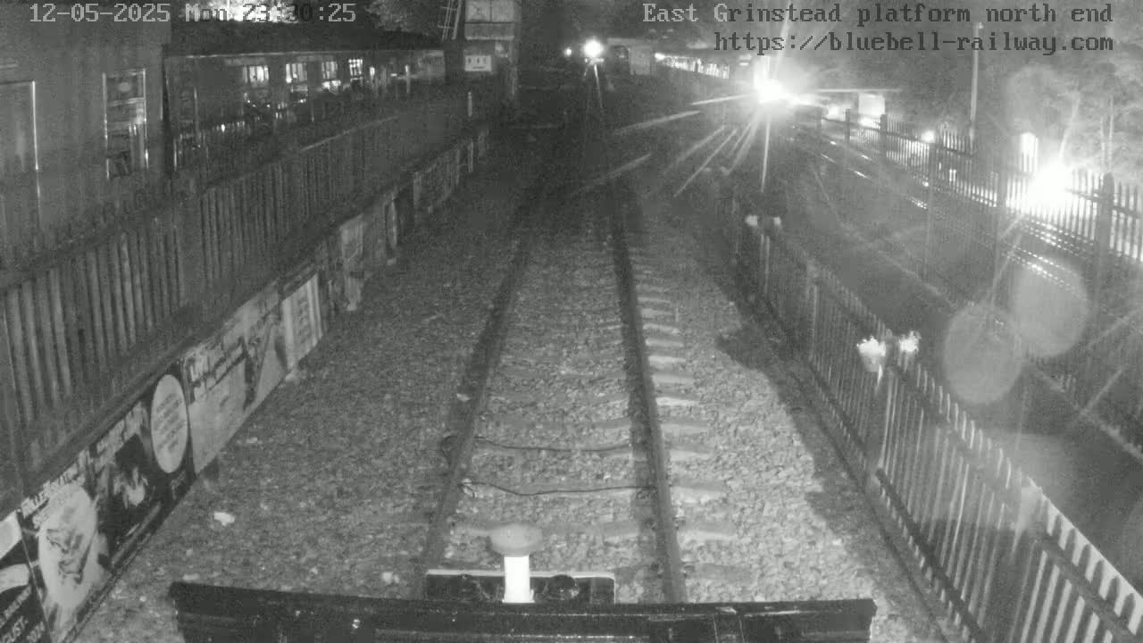 A nighttime view of railway tracks at a station platform, bordered by fences and posters, under dark conditions.