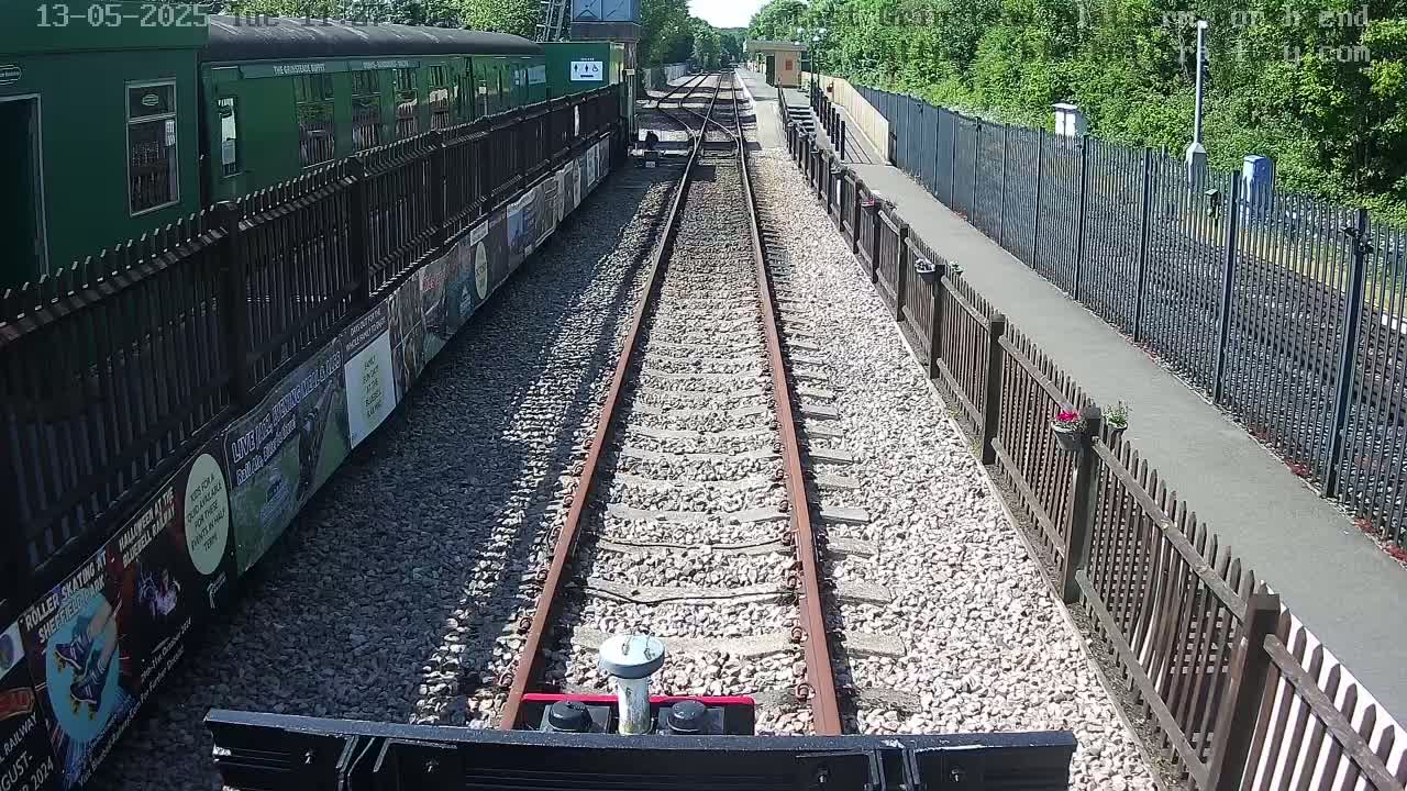 A train's-eye view shows a railway track on a sunny day, bordered by fences and a green train car on one side, with a walking path on the other.