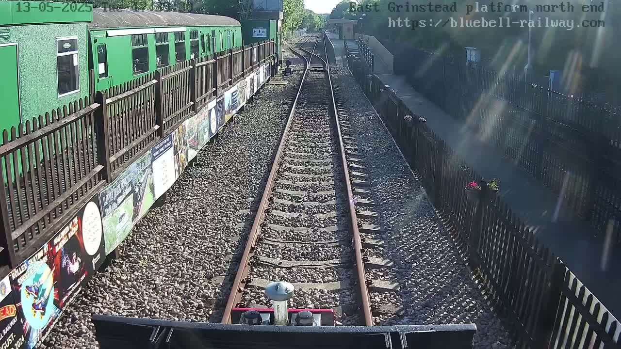 A view from a train shows a sunny day at a railway station with a green train alongside the tracks and a gravel path between the tracks and a black fence.