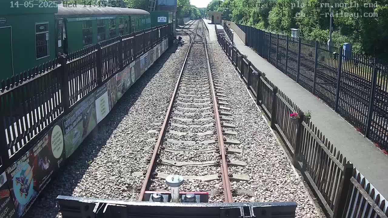 A train travels down a sunny railway track between wooden fences, with a green train car visible to the side.