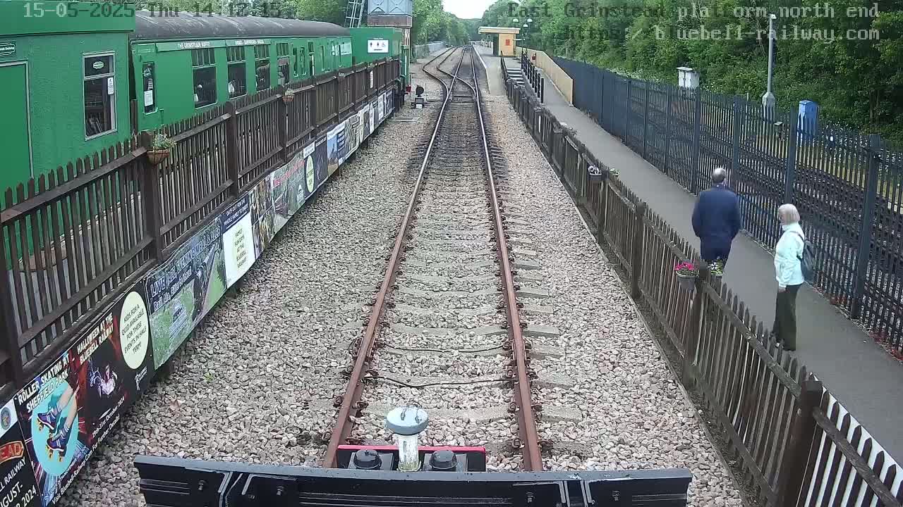 A view from a train shows a railway track extending to a station platform where two people are walking, alongside a green train and wooden fences.