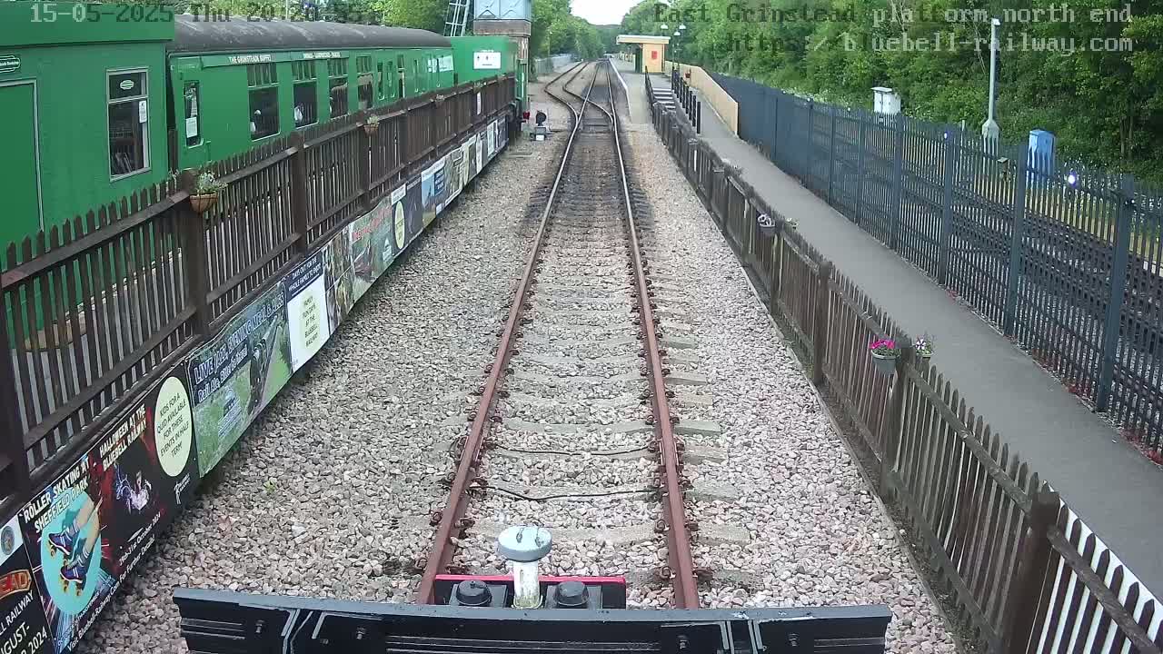 A train's-eye view of railway tracks extending to a station platform bordered by wooden fences, with a green train car visible to the left, under overcast conditions.