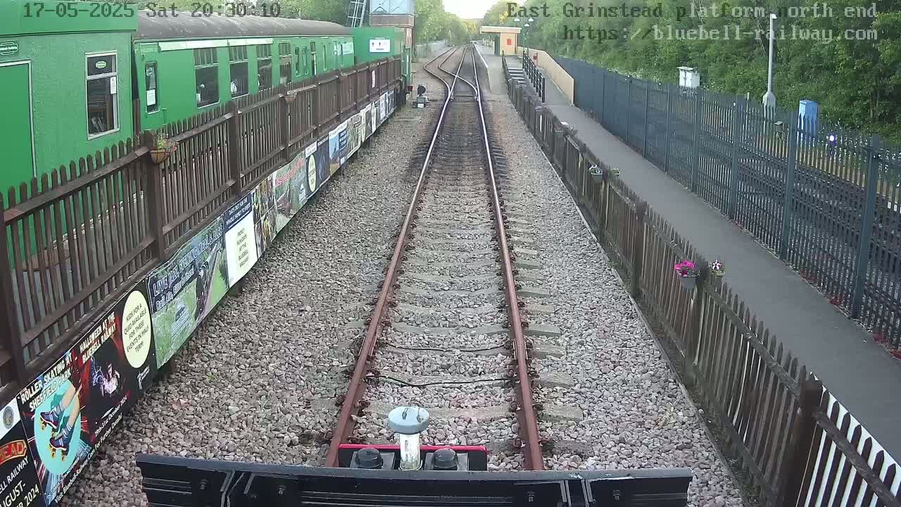 A view from a train's perspective shows railway tracks leading to a station platform, bordered by fences and lined with posters, under the cover of evening darkness.