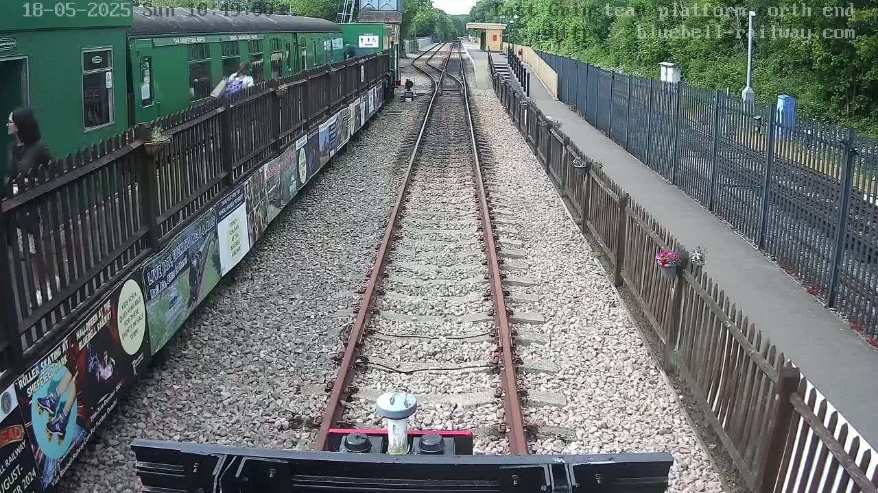 A train track stretches into the distance between wooden fences, with a green train car visible to the left and a sunny day.