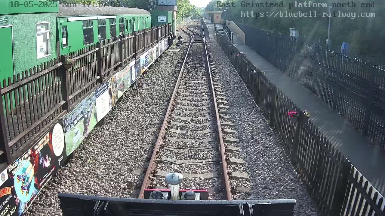 A view from a train's perspective shows a railway track leading to a station platform on a sunny day, with a green train alongside the tracks and a fence separating the platform from a path.
