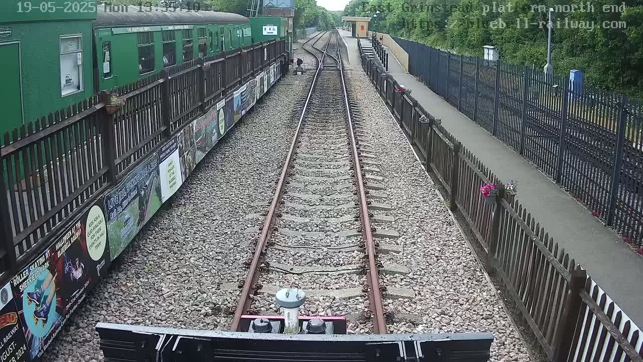 A view from a train shows a railway track extending into the distance between wooden fences, alongside a green train car and a small station platform, on a sunny day.