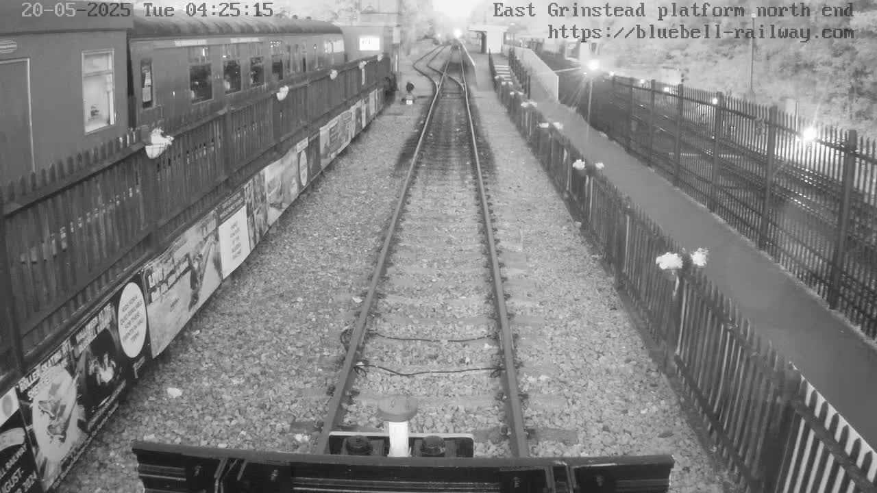 A nighttime view of a train platform with a train alongside the tracks, fences lining the platform, and gravel between the rails.