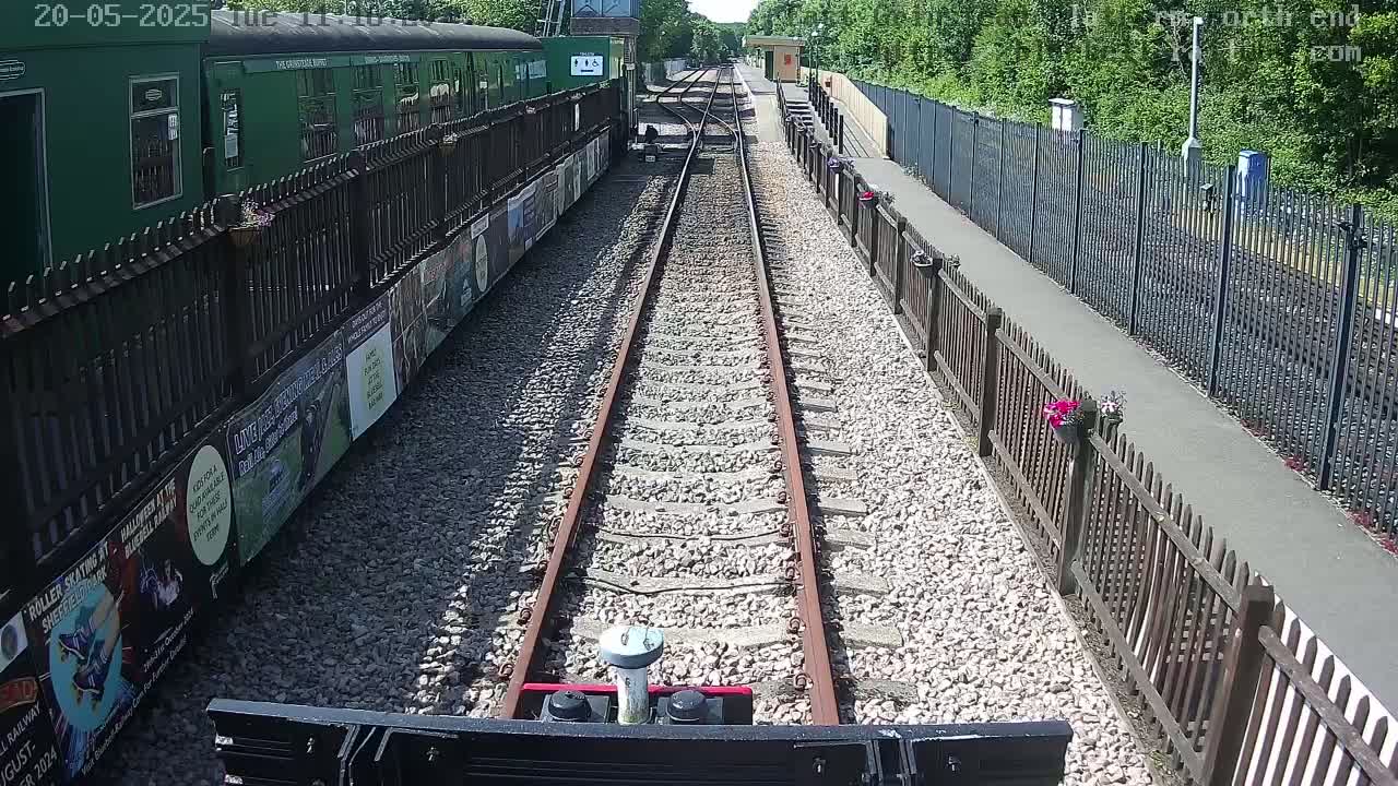 A railway track extends into the distance between wooden fences, alongside a green train car and a paved walkway on a sunny day.