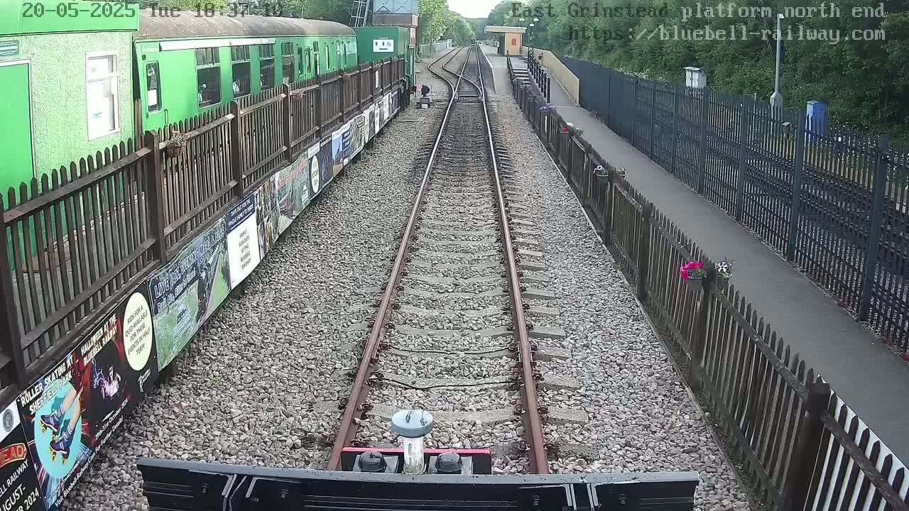 A view from a train down a railway track lined with gravel, fences, and posters, on an overcast day.