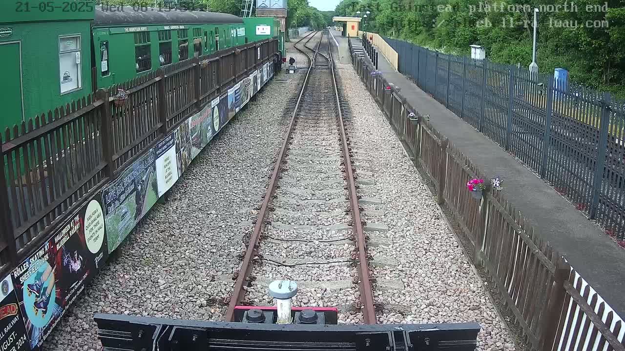 A view from a train on a railway track, with a green train car and posters visible to the side, bordered by fences, under a partly sunny sky.