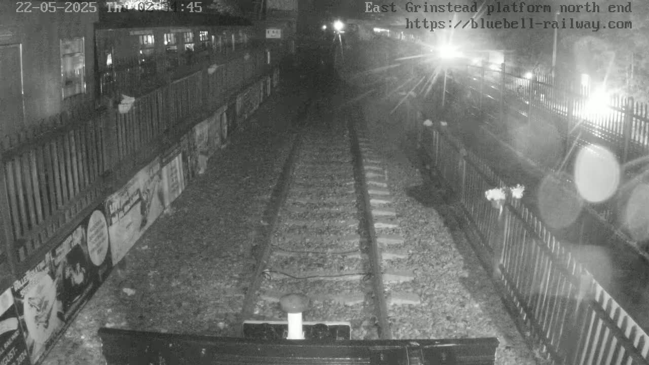 A nighttime view of train tracks at a platform, bordered by fences and posters, under artificial light.