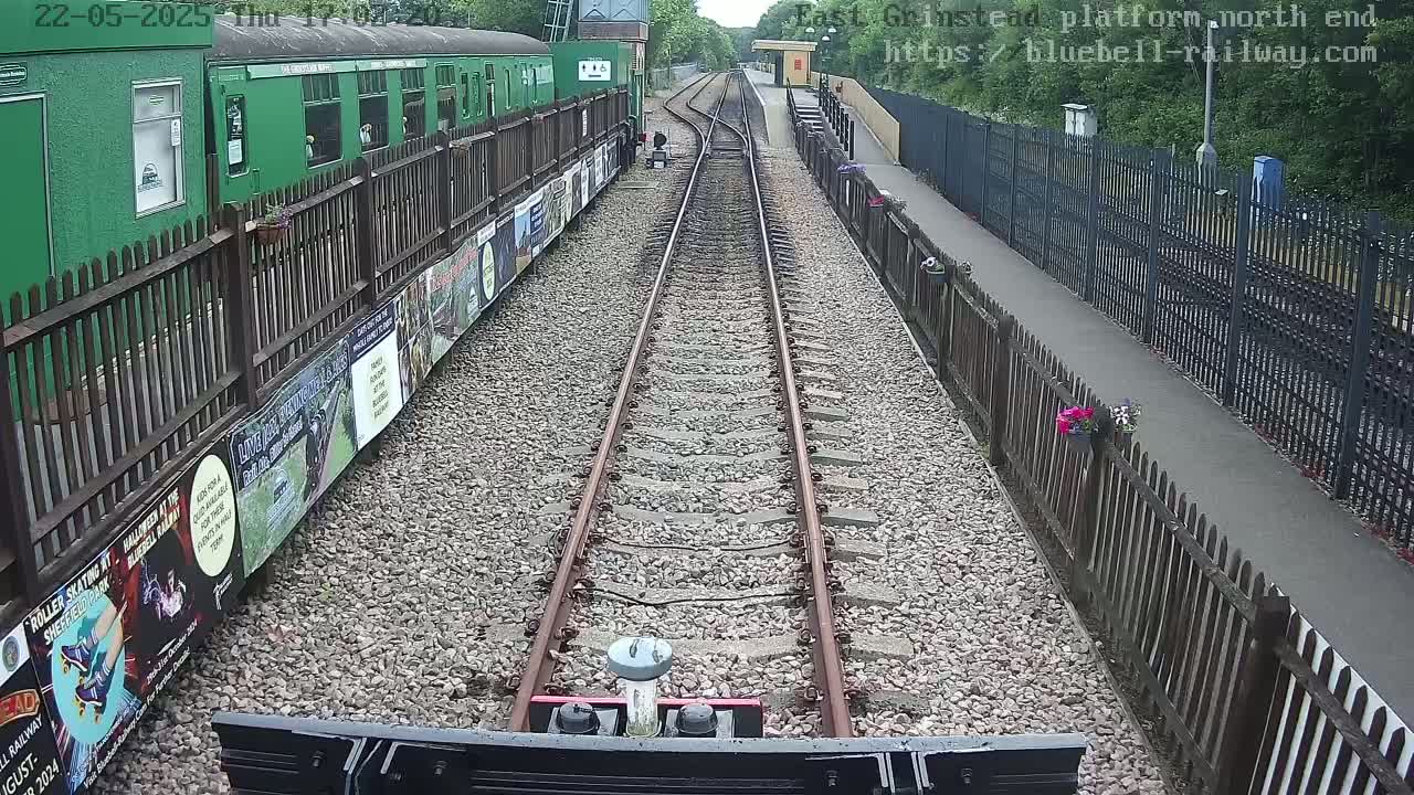 A view from a train's cab shows a railway track stretching toward a station platform bordered by fences, with a green train car to the left and lush greenery beyond.