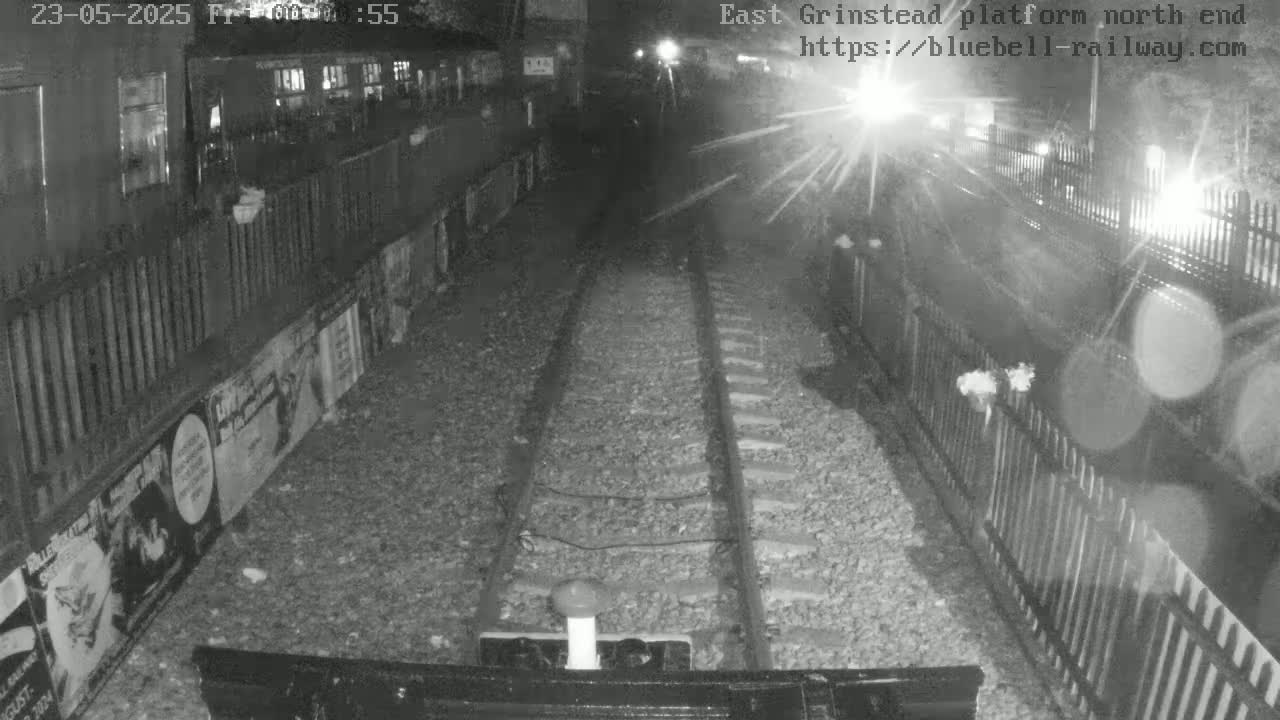 A nighttime, low-light view from a train shows railway tracks extending into the distance, bordered by fences and posters on a wall.