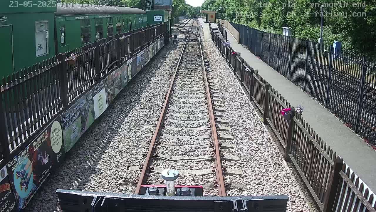 A view from a train down a railway track on a sunny day, with a green train car and wooden fences along the platform.