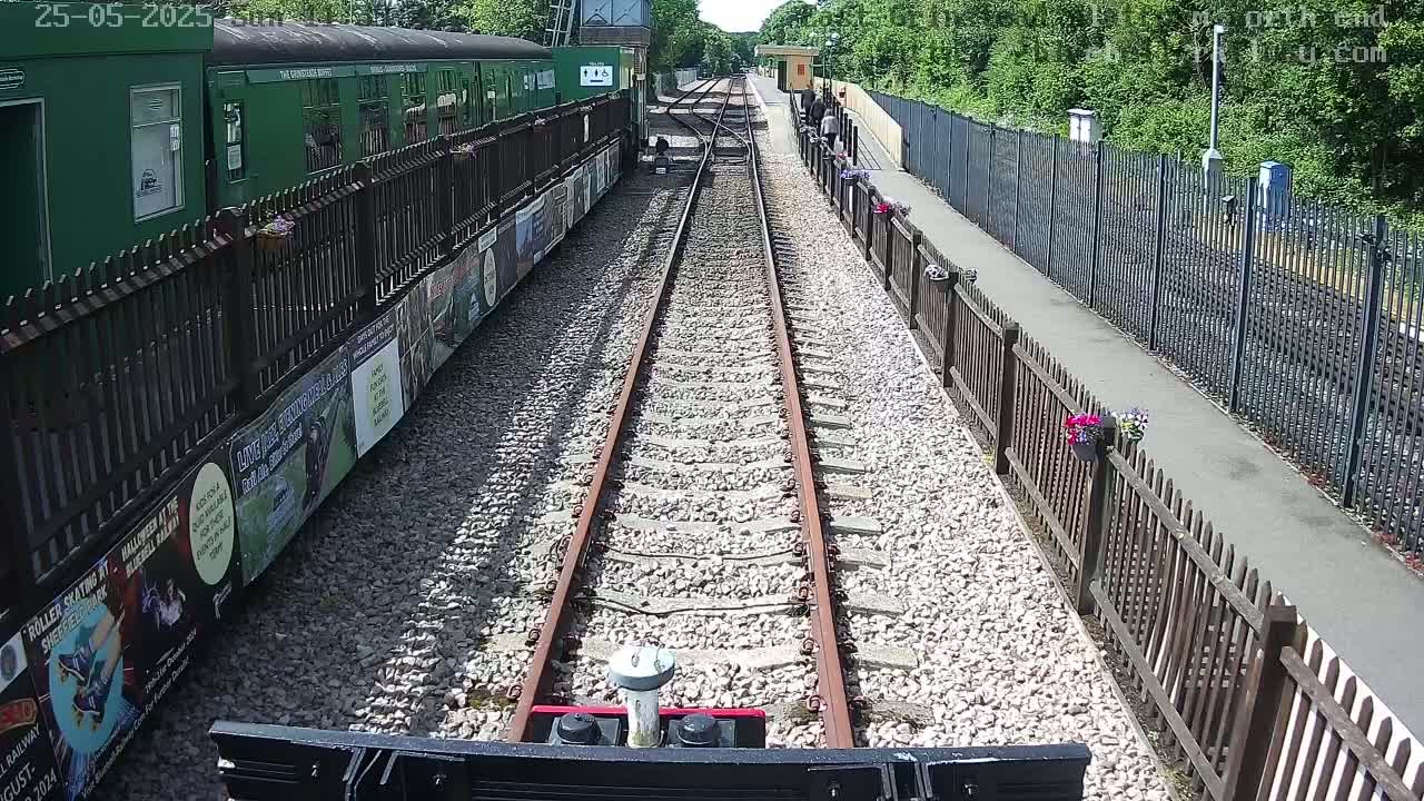 A view from a train shows a railway track extending into the distance between wooden fences, lined with posters and flowerpots, on a sunny day.