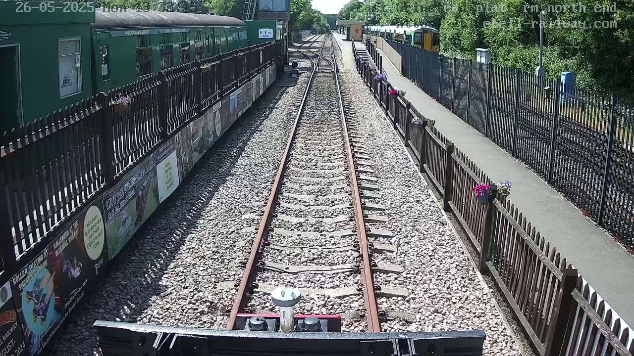 A view from a train's perspective down a railway track on a sunny day, with green train carriages and wooden fences on either side.