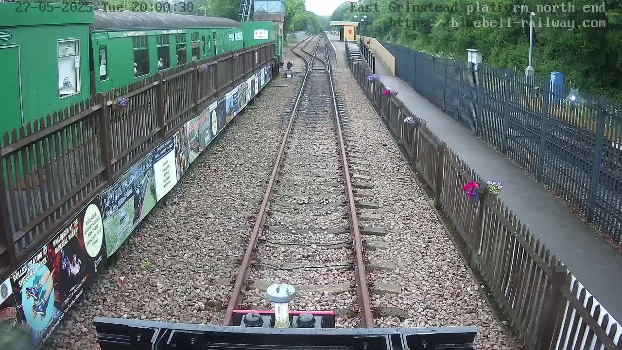 A train's-eye view of a railway track leading to a platform with a green train car and wooden fences lined with flowers, under overcast skies.