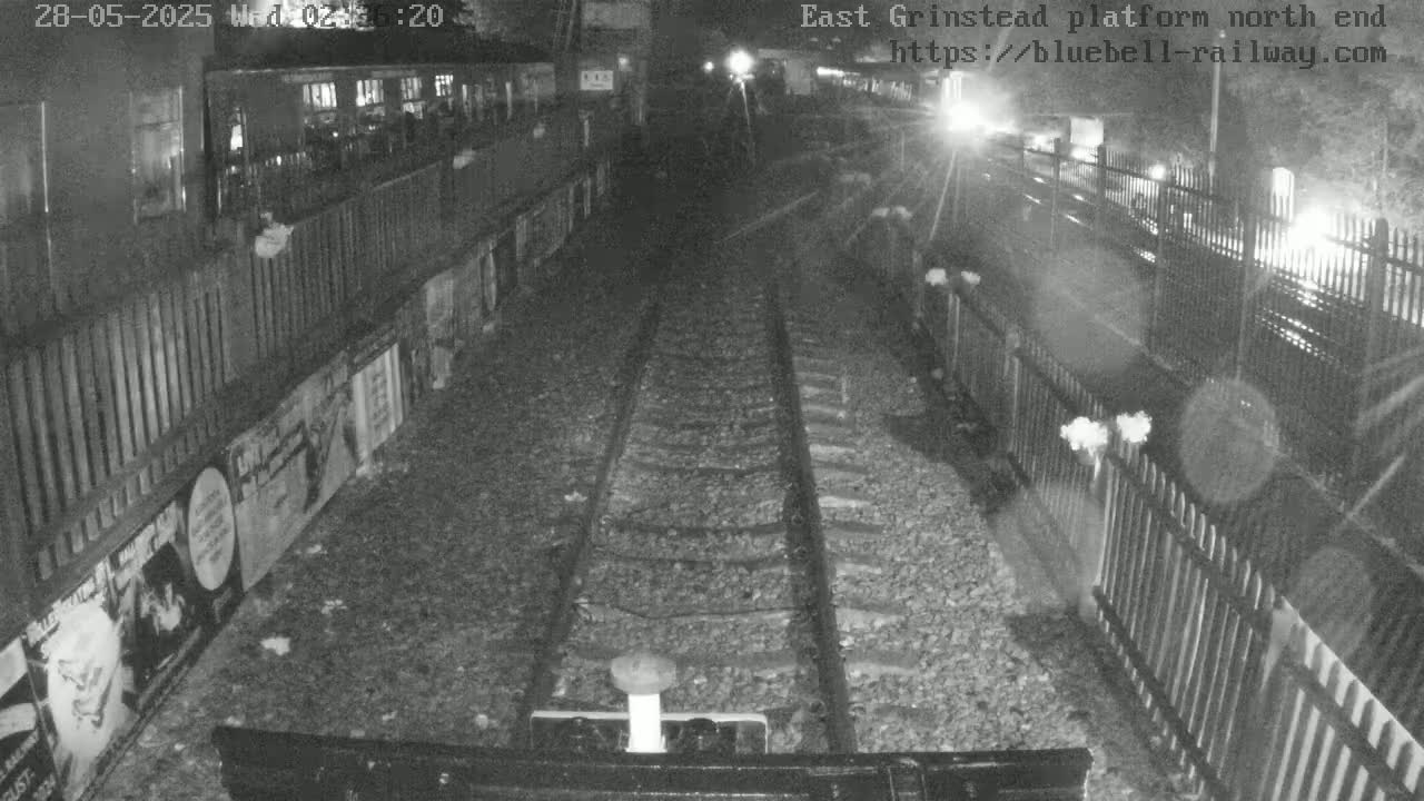 A nighttime view of train tracks at a station platform, with fences and advertisements visible along the sides.