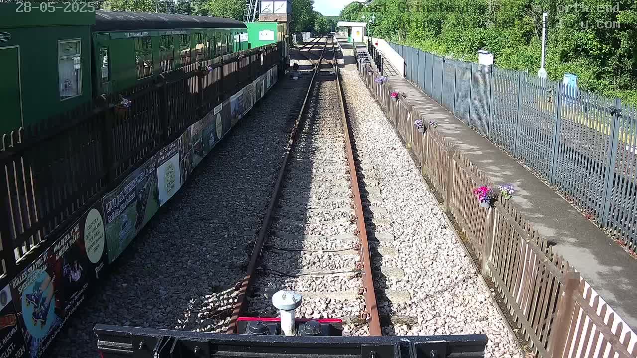 A train sits at a sunny station with flower boxes lining the platform and a view down a railway track.