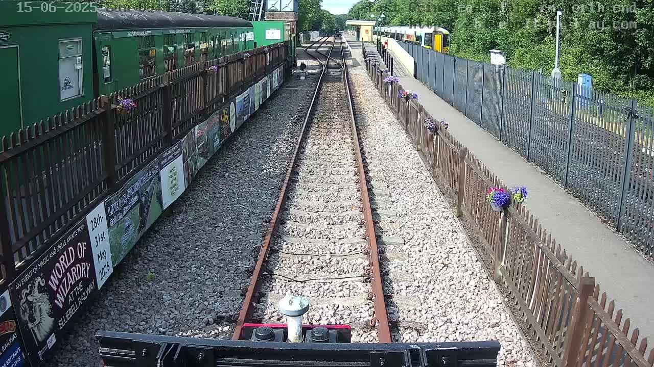 A train's front-facing view shows dual railway tracks on a sunny day, bordered by wooden fences with flower pots and posters along the platform.