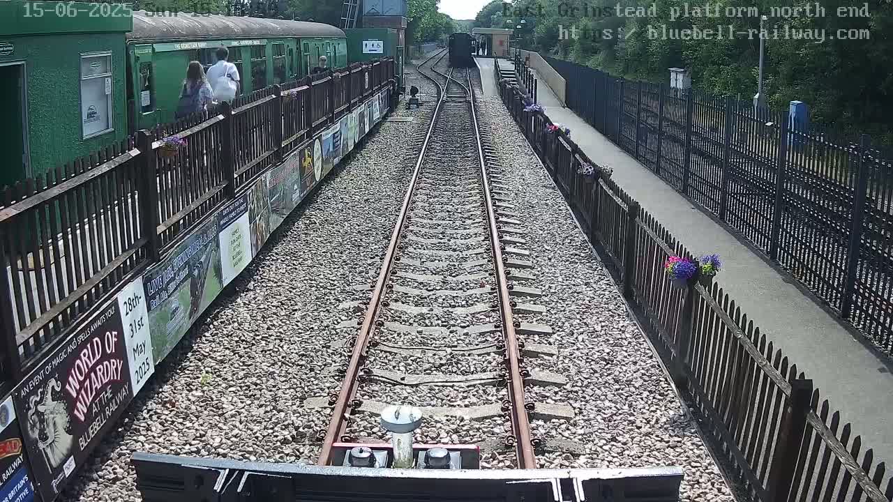 A train's-eye view shows a sunny day at a railway station with a green train and passengers on a platform, alongside a gravel path with fences and flowerpots.
