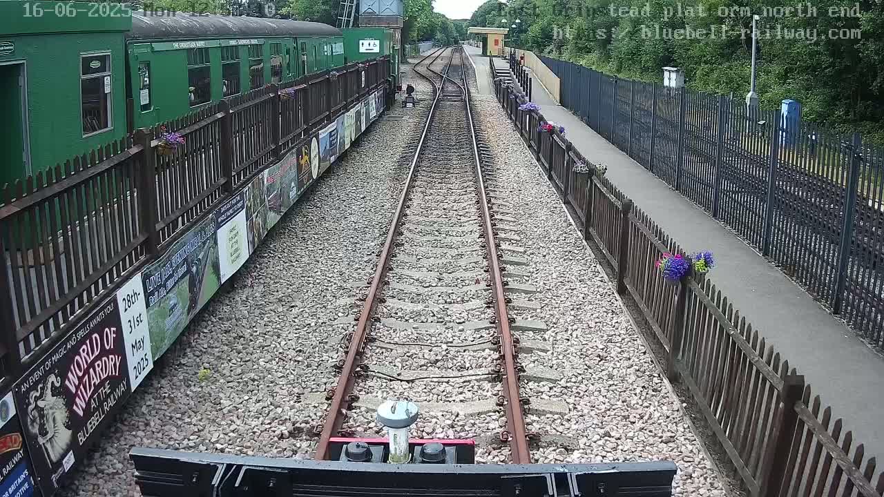 A train's view down a railway track on a sunny day, with a green train car and platform visible to the left, and wooden fences and a paved walkway on the right.