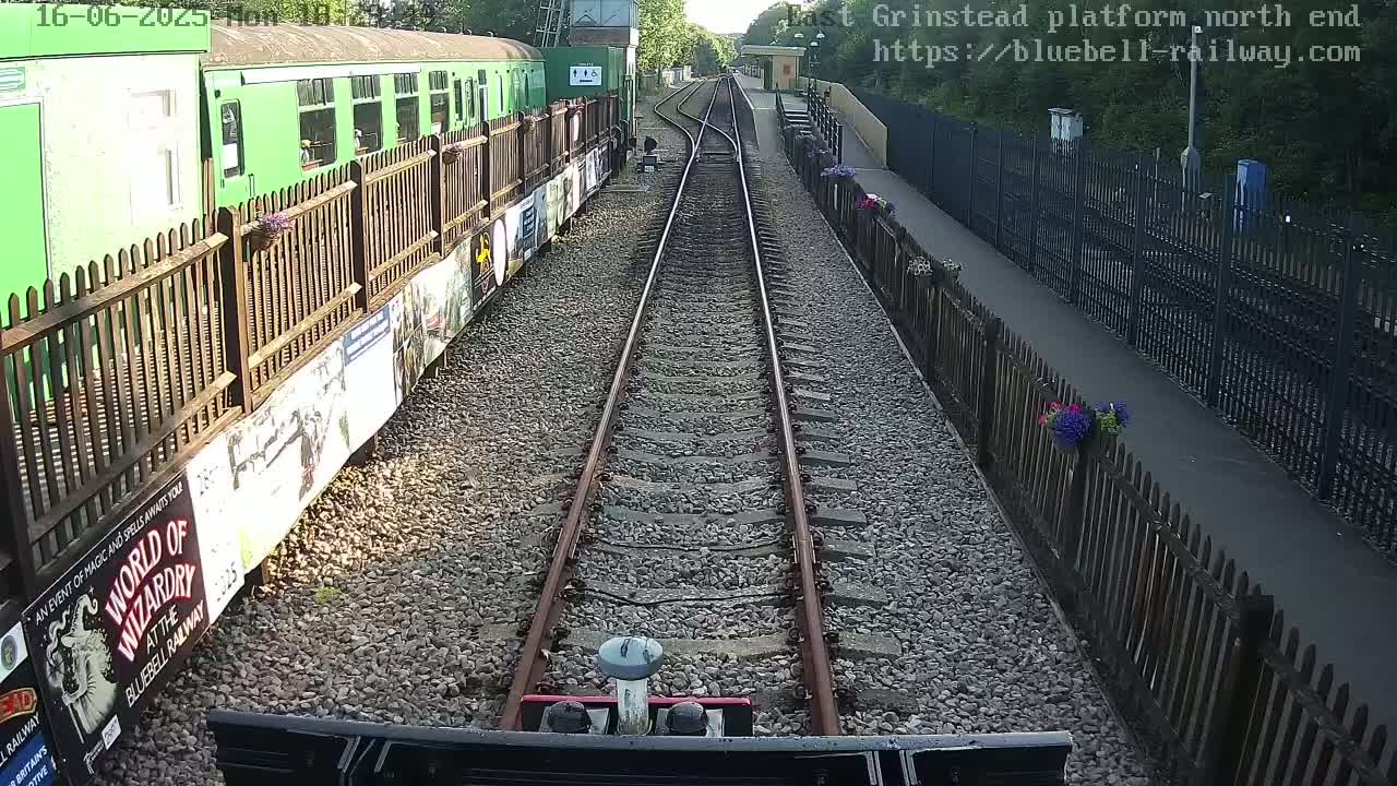 A view from a train shows two parallel train tracks extending to a station platform bordered by wooden fences and flower boxes on a sunny day.