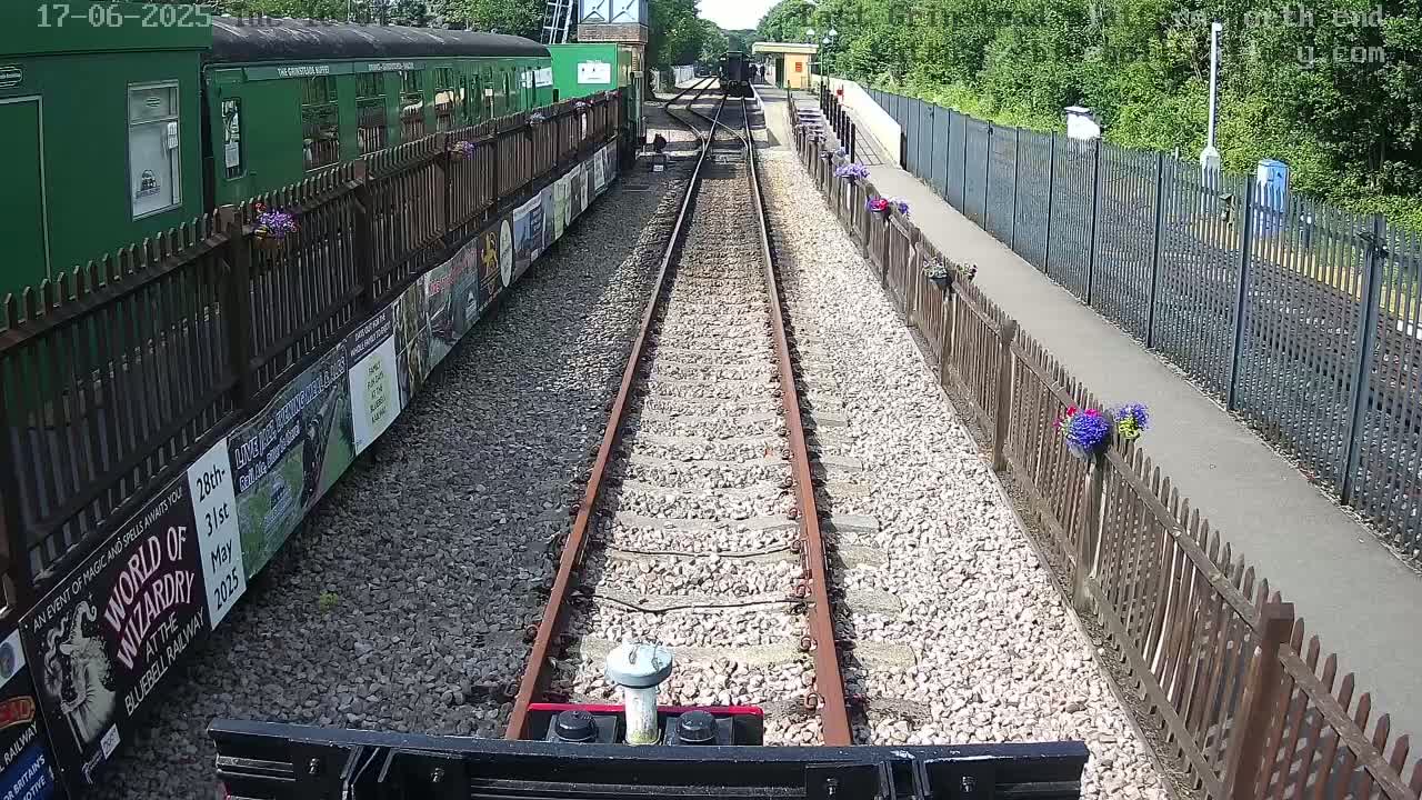 A train travels down a railway track on a sunny day, passing between wooden fences lined with flower boxes and posters.