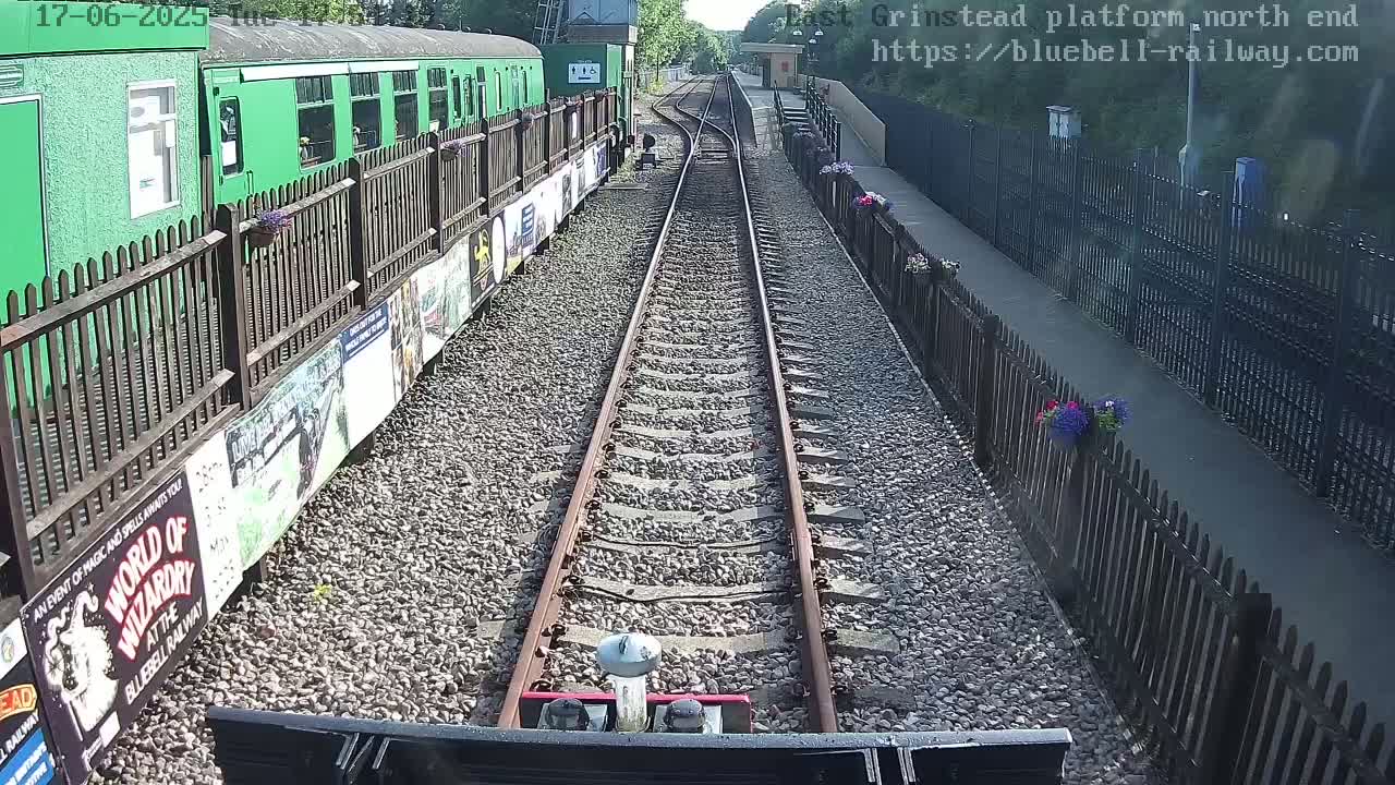 A train's-eye view shows a railway track extending into the distance on a sunny day, alongside a green train and platform with wooden fences and flower pots.
