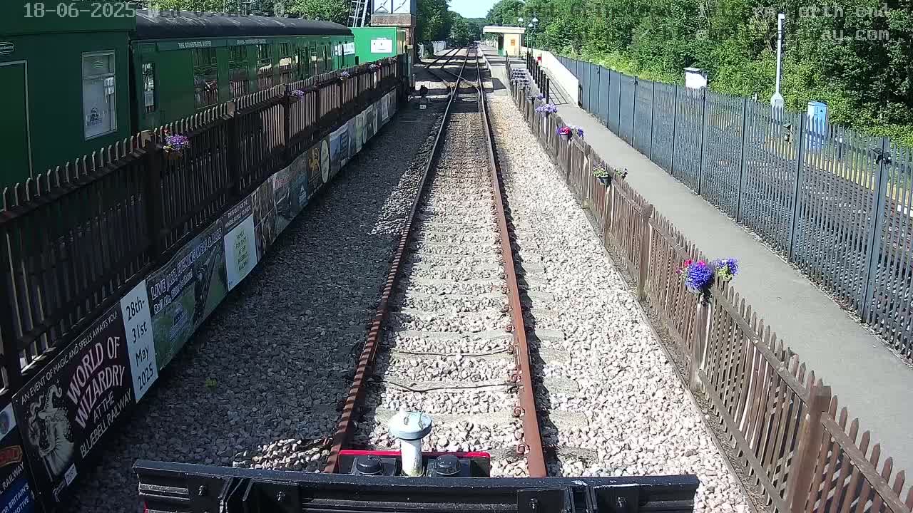 A train’s point of view shows a sunny day at a railway station with a gravel path, wooden fences, and flower pots lining the sides of the tracks.