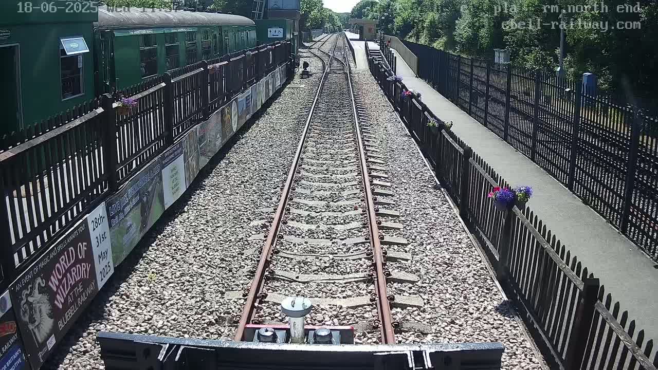 A view from a train shows a sunny day at a railway station with a green passenger car and a gravel path alongside the tracks, bordered by black fences and small flower pots.