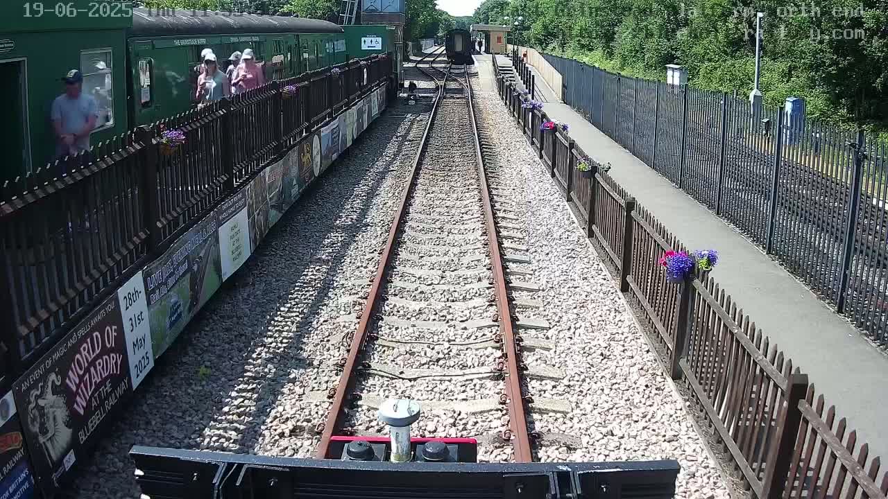 A train's view of a railway track on a sunny day, with people standing near a train at a station lined with fences and flower pots.