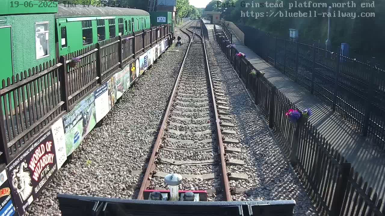 A view from a train shows a railway track extending into the distance, bordered by fences with flower planters and posters, on a sunny day.