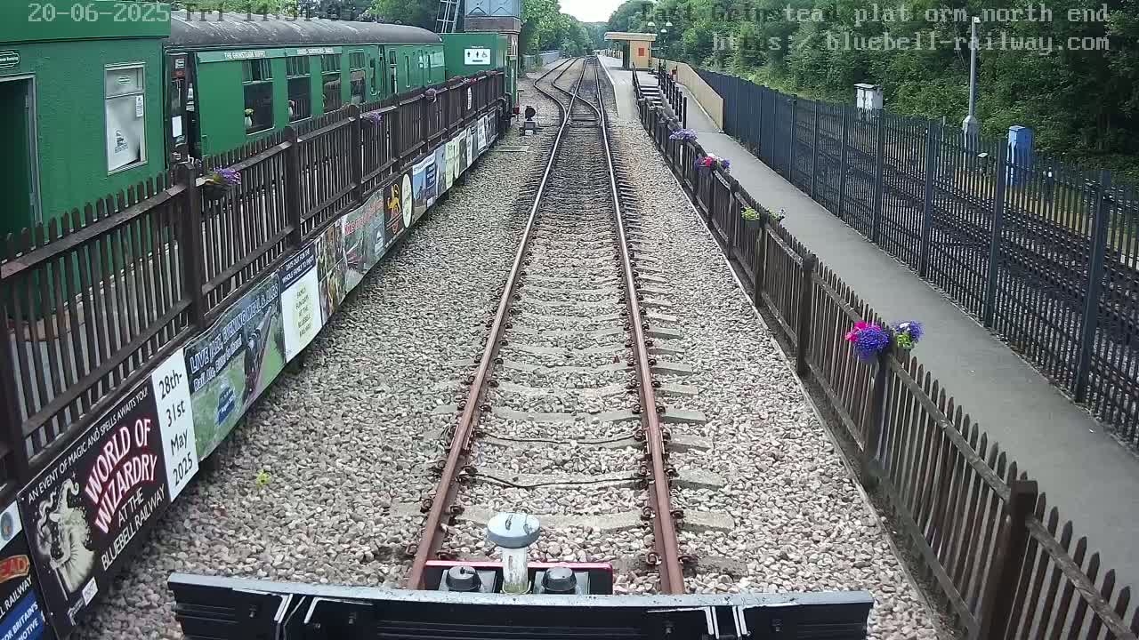 A train travels down a railway track between wooden fences, lined with posters, on a sunny day.