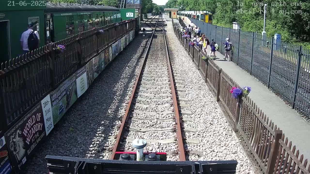 A train approaches a station on a sunny day, with people standing on the platform and a green train parked nearby.