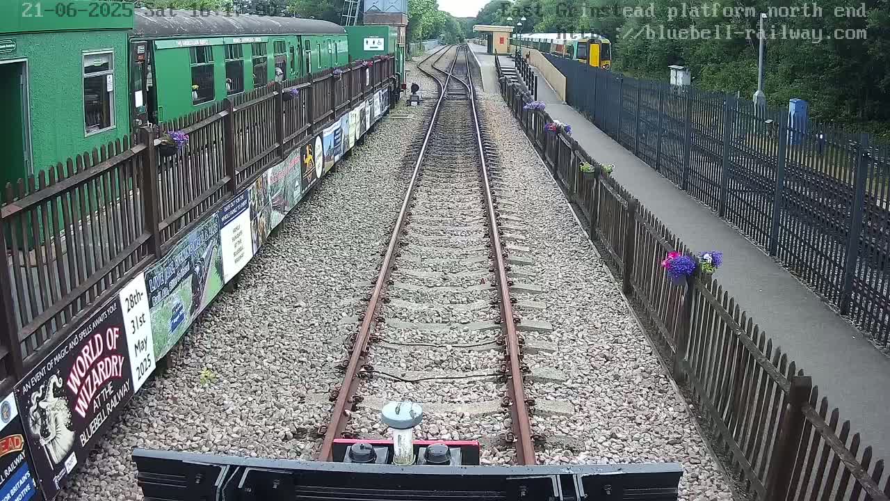 A view from a train shows railway tracks leading to a station platform bordered by fences and flower boxes, on a sunny day.