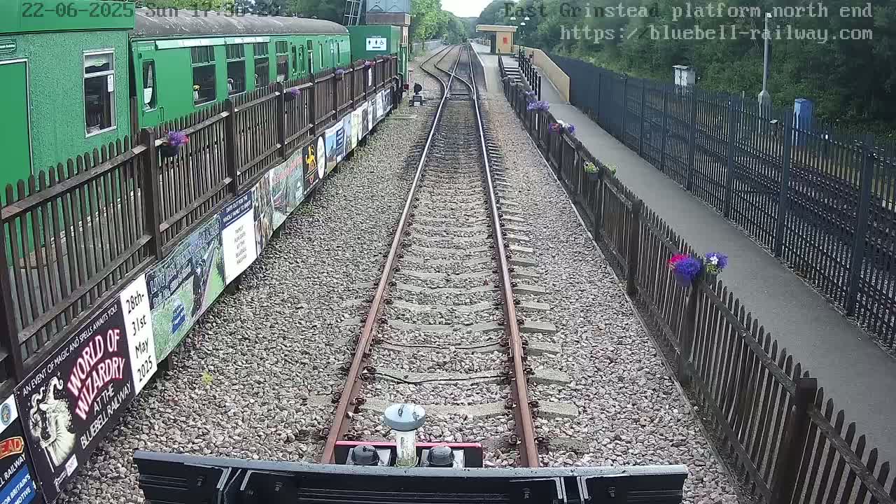 A view down a railway track on a sunny day, showing a green train and platform with flower boxes along the fences.