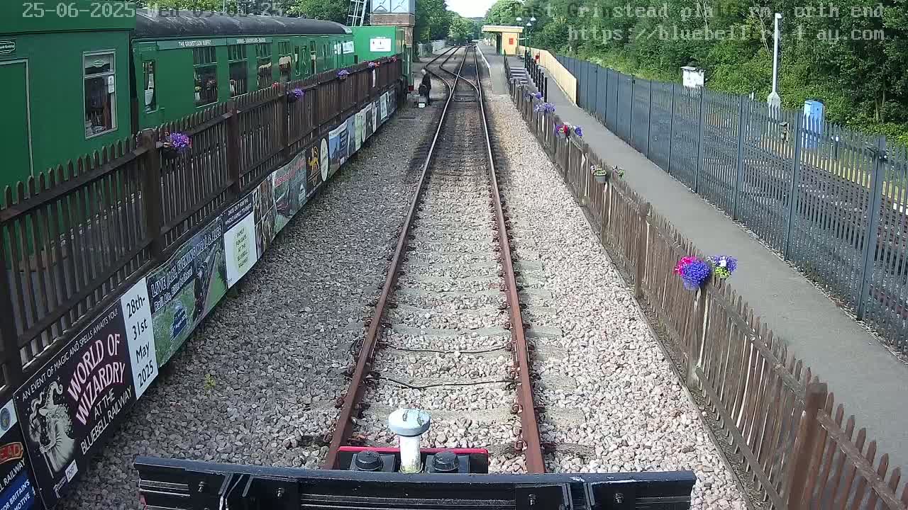 A view from a train's perspective shows a railway track lined with a green train car and fences with flowers, under a sunny sky.